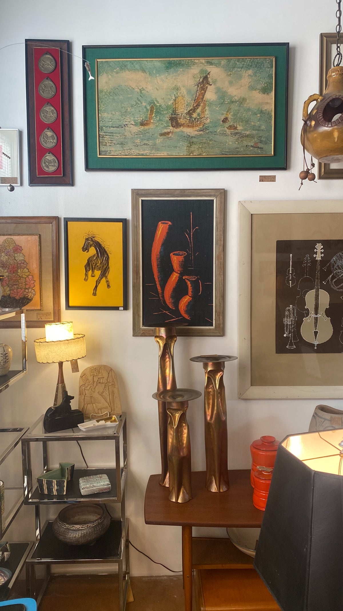 A collection of framed art prints and decorative objects displayed on a white wall in a shop or gallery setting, including a central vintage-style painting of red vases, a yellow horse print, and a green sea scene.